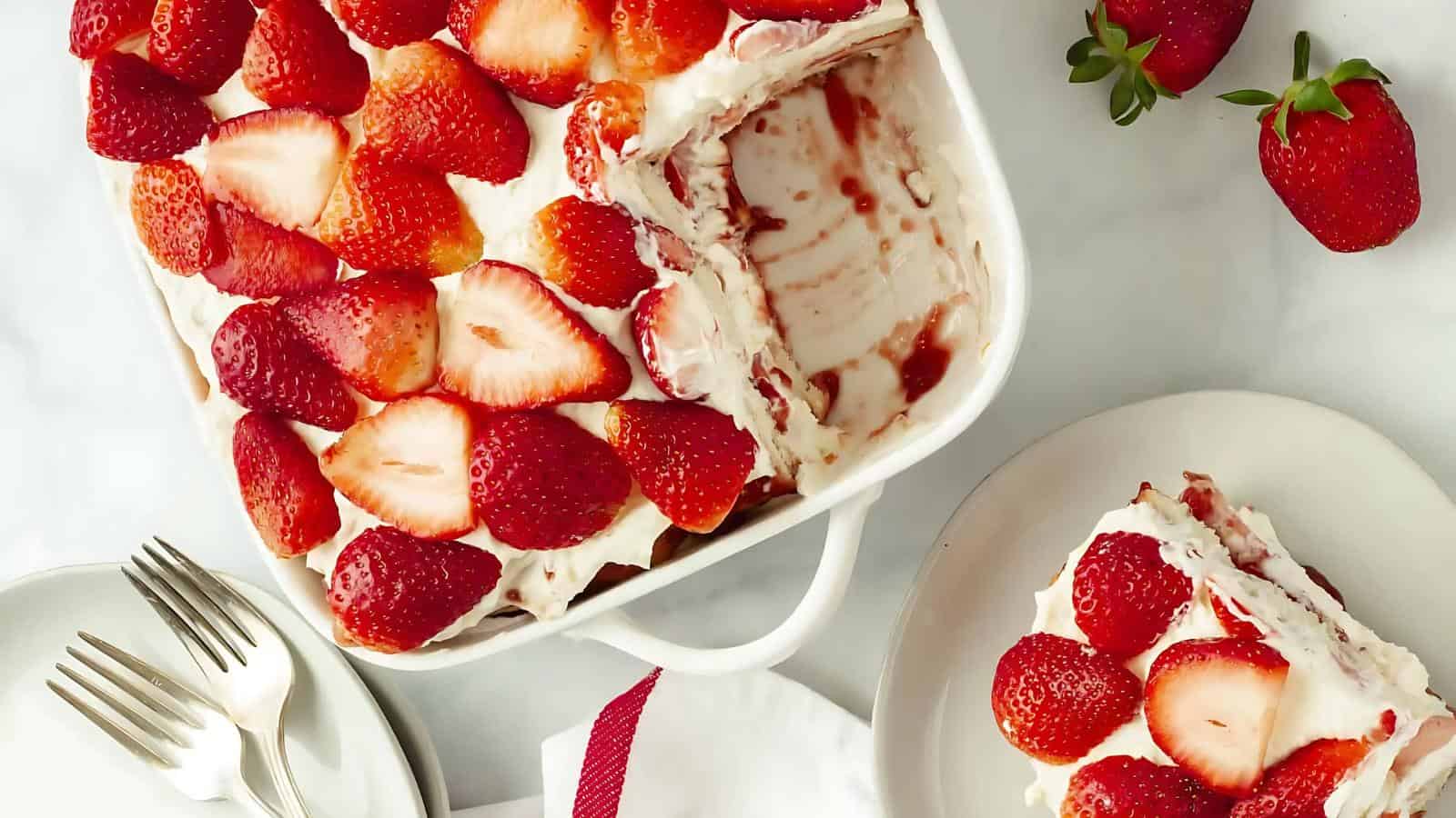 A baking dish with sliced strawberries and cream dessert, a portion served on a plate beside it, with fresh strawberries and forks nearby.