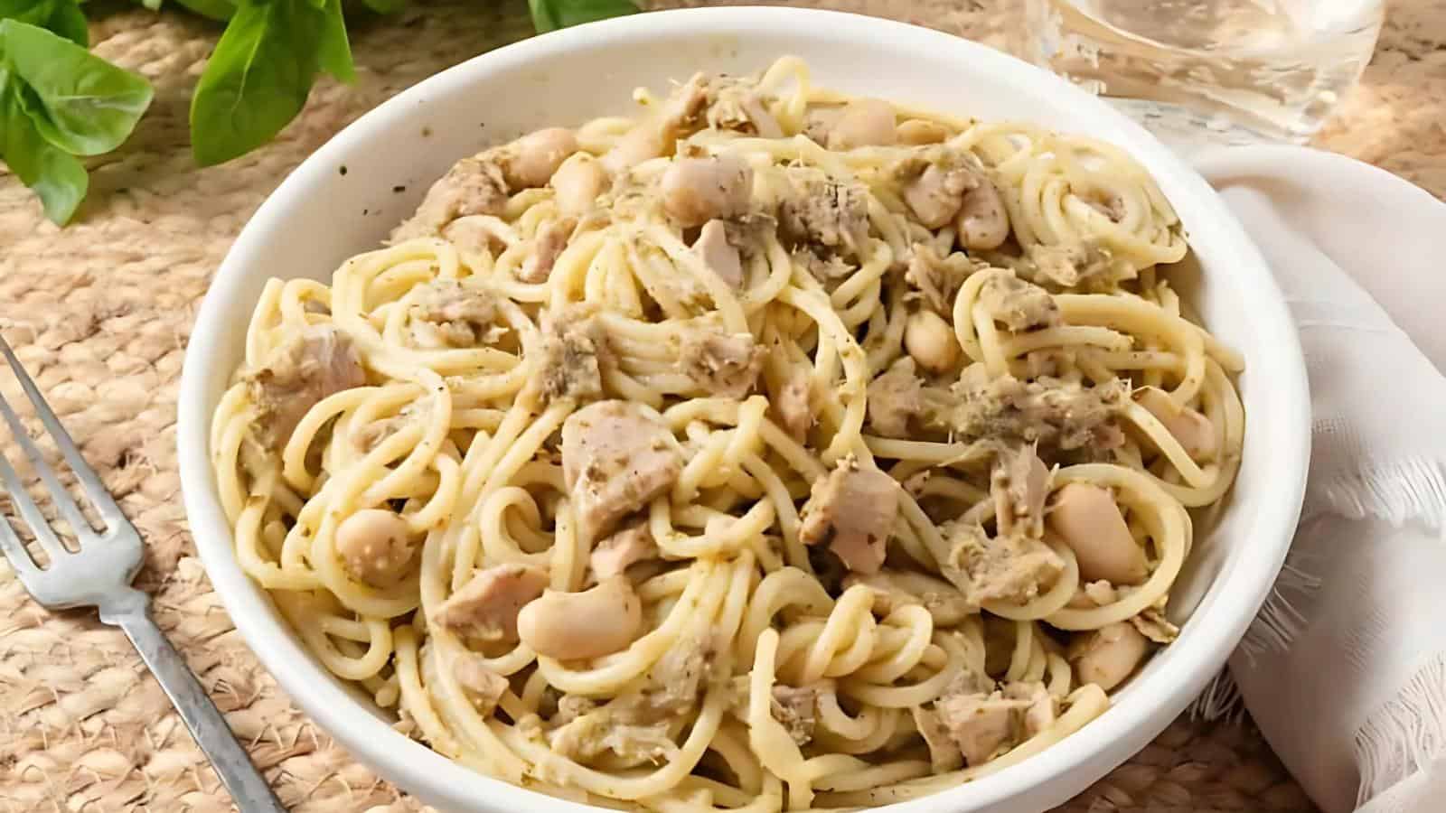 A white bowl of spaghetti mixed with chunks of chicken, white beans, and a green herb sauce sits on a woven placemat beside a fork, napkin, and glass of water.