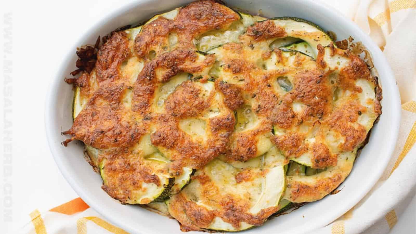 A baked zucchini casserole in a white oval dish, topped with a golden, melted cheese crust, sits on a white and yellow cloth.