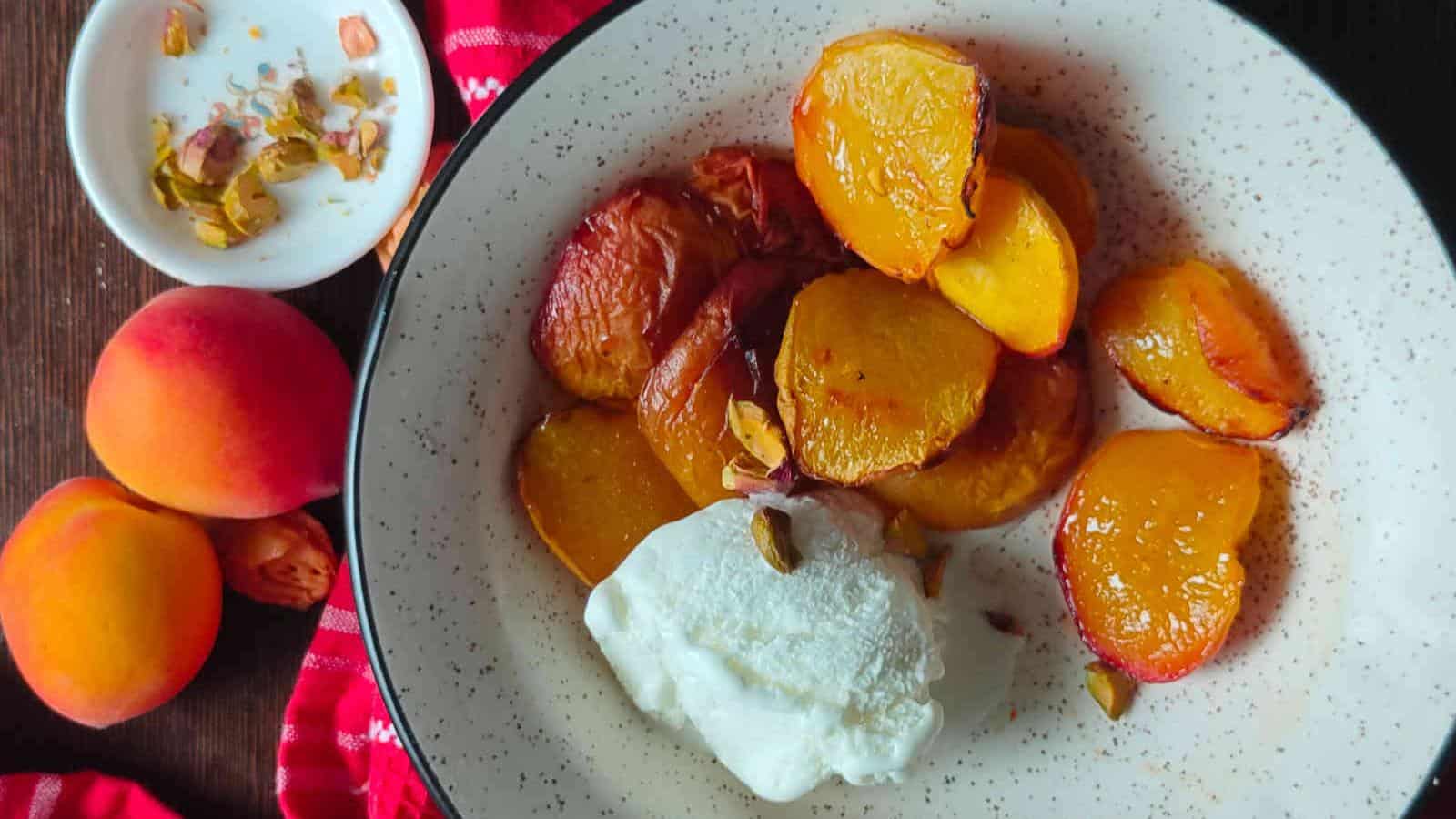 A bowl with roasted peach slices, a scoop of white cream or yogurt, and chopped nuts; whole peaches and a small dish of nuts are beside the bowl on a dark table.
