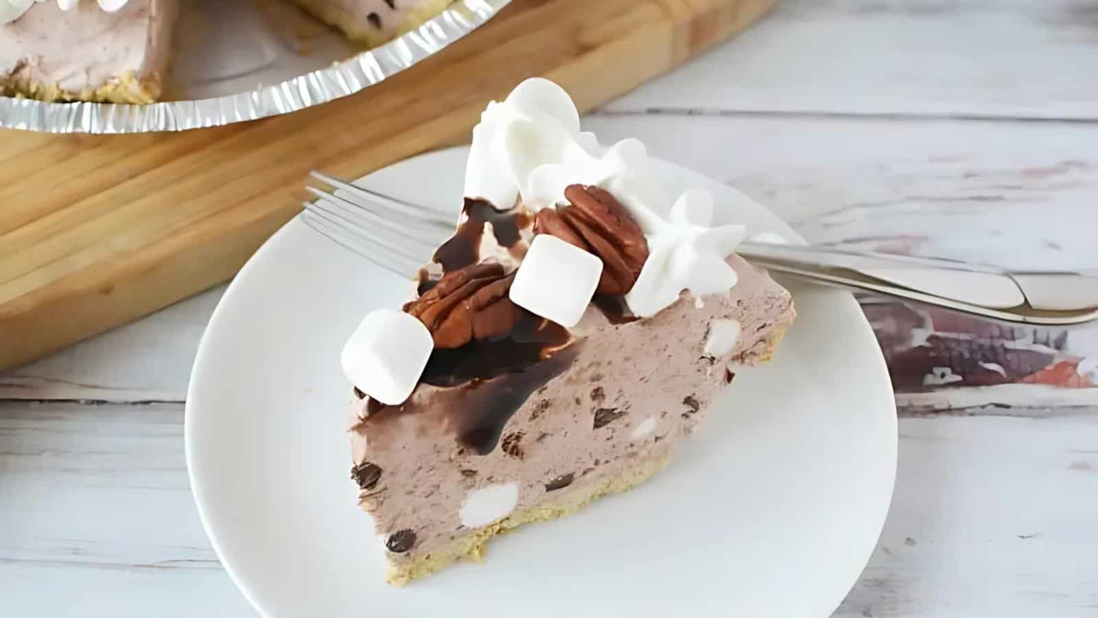A slice of chocolate chip and marshmallow pie on a white plate, topped with whipped cream, chocolate syrup, pecan halves, and marshmallows, with a fork beside it.