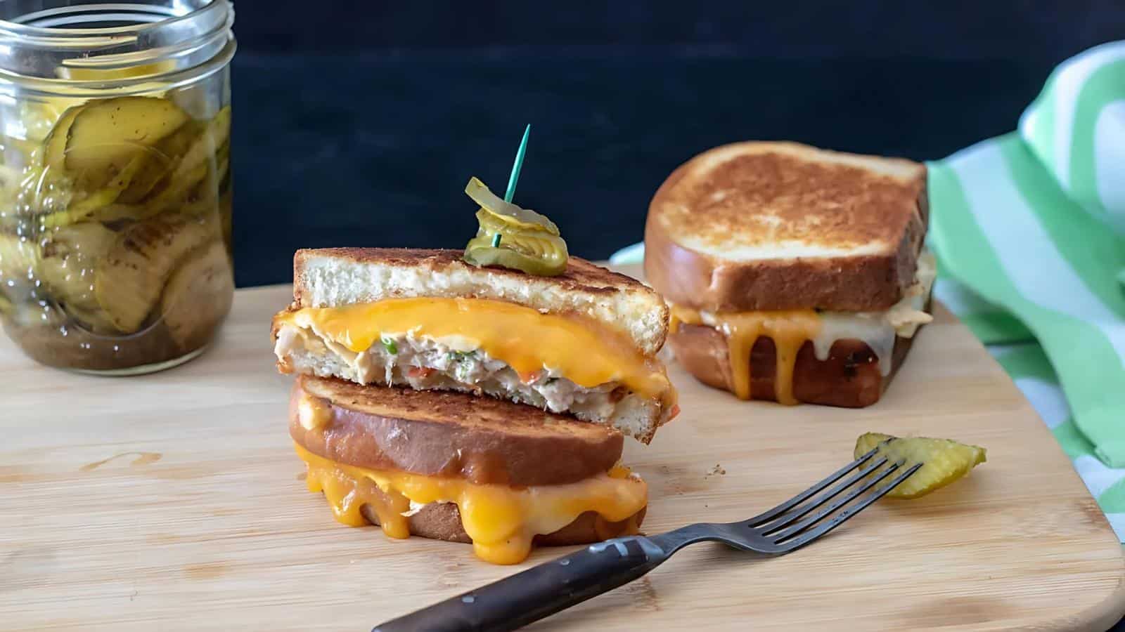 Two grilled cheese sandwiches with melted cheddar on a wooden board, one cut in half and topped with pickles, with a fork and a jar of pickles beside them.