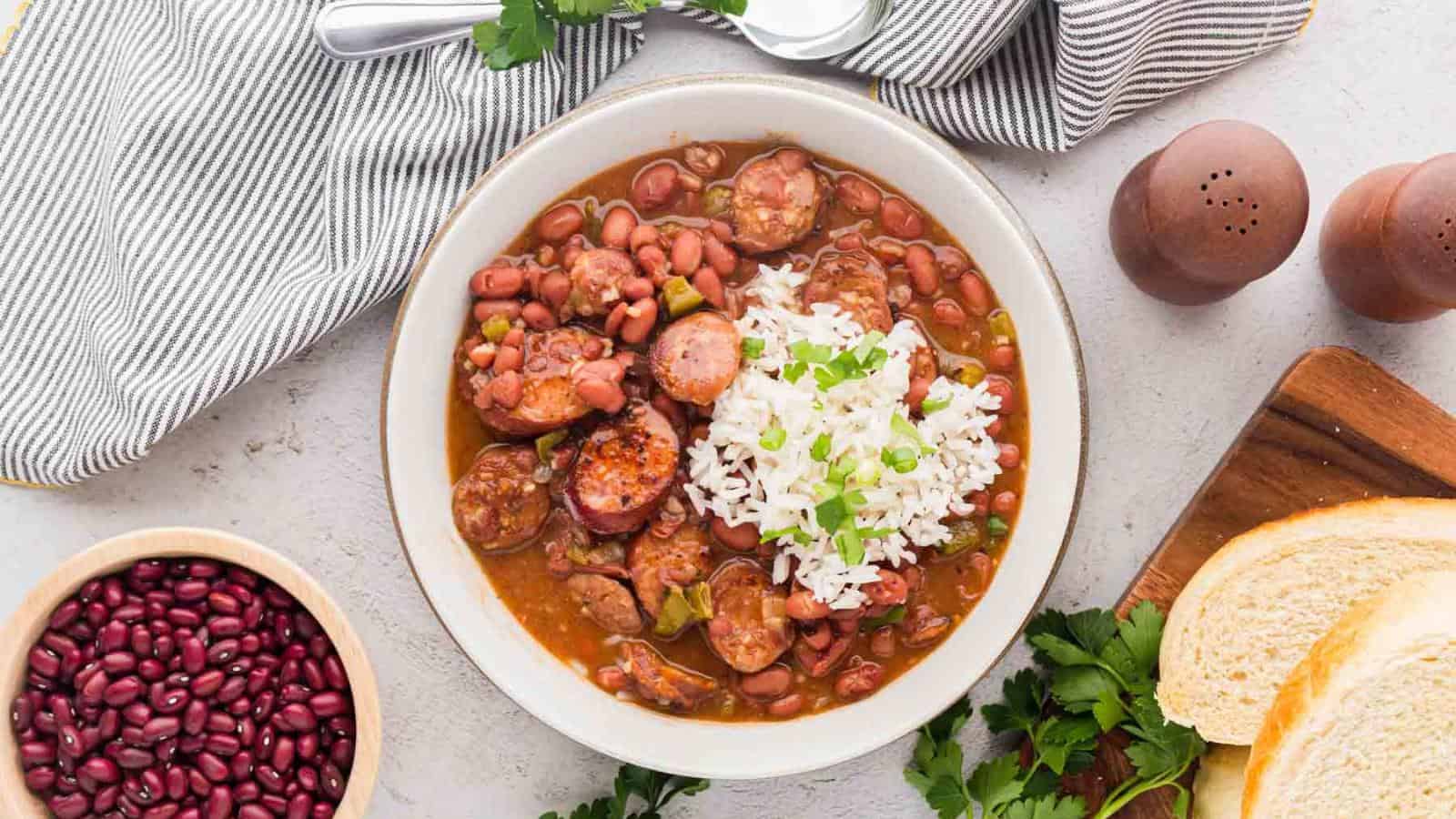 A bowl of red beans and sausage stew topped with white rice and green onions, surrounded by bread, herbs, red beans, salt and pepper shakers, and a striped napkin with a spoon.