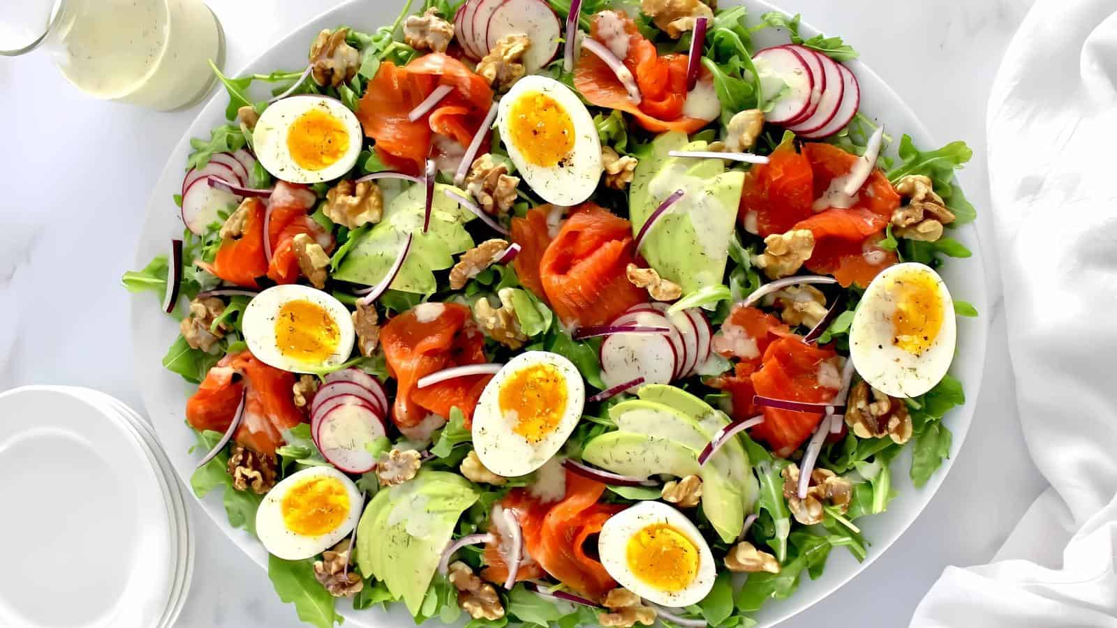 A salad with arugula, sliced avocado, soft-boiled eggs, smoked salmon, radishes, walnuts, and red onion is arranged on a white platter with a dressing on the side.