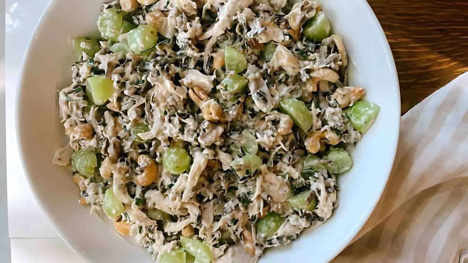 A white bowl filled with a mixture of shredded chicken, wild rice, green grapes, cashews, and herbs, coated in a creamy dressing.
