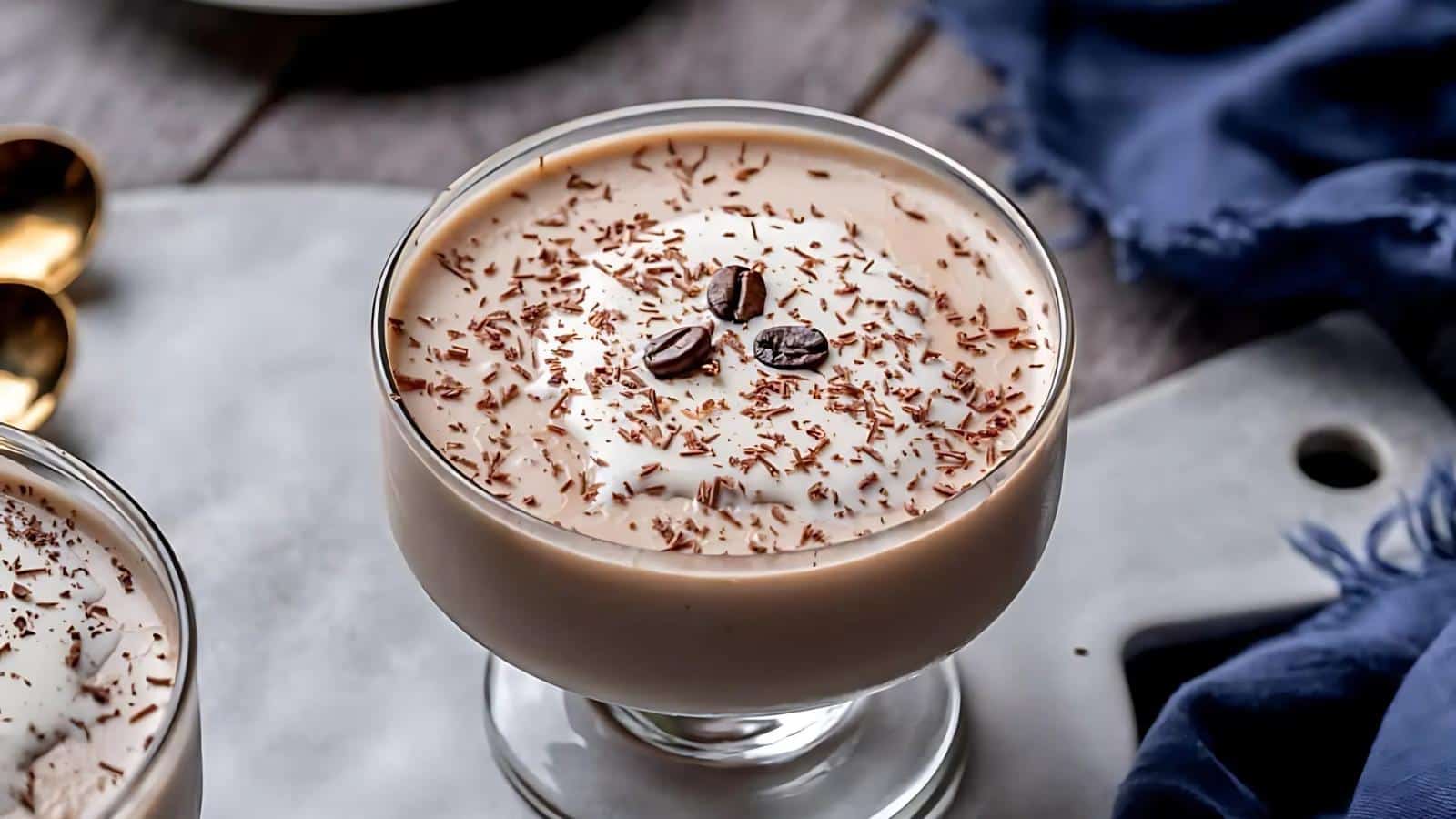 A glass dessert dish filled with creamy mousse, topped with chocolate shavings and three coffee beans, placed on a marble surface.