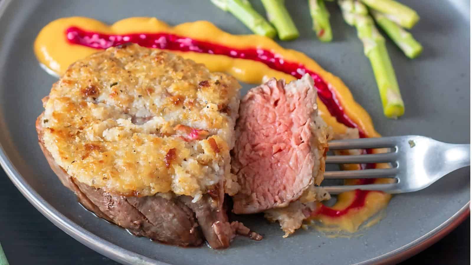 A slice of stuffed beef tenderloin on a gray plate with a fork, accompanied by asparagus, yellow sauce, and a red sauce drizzle.