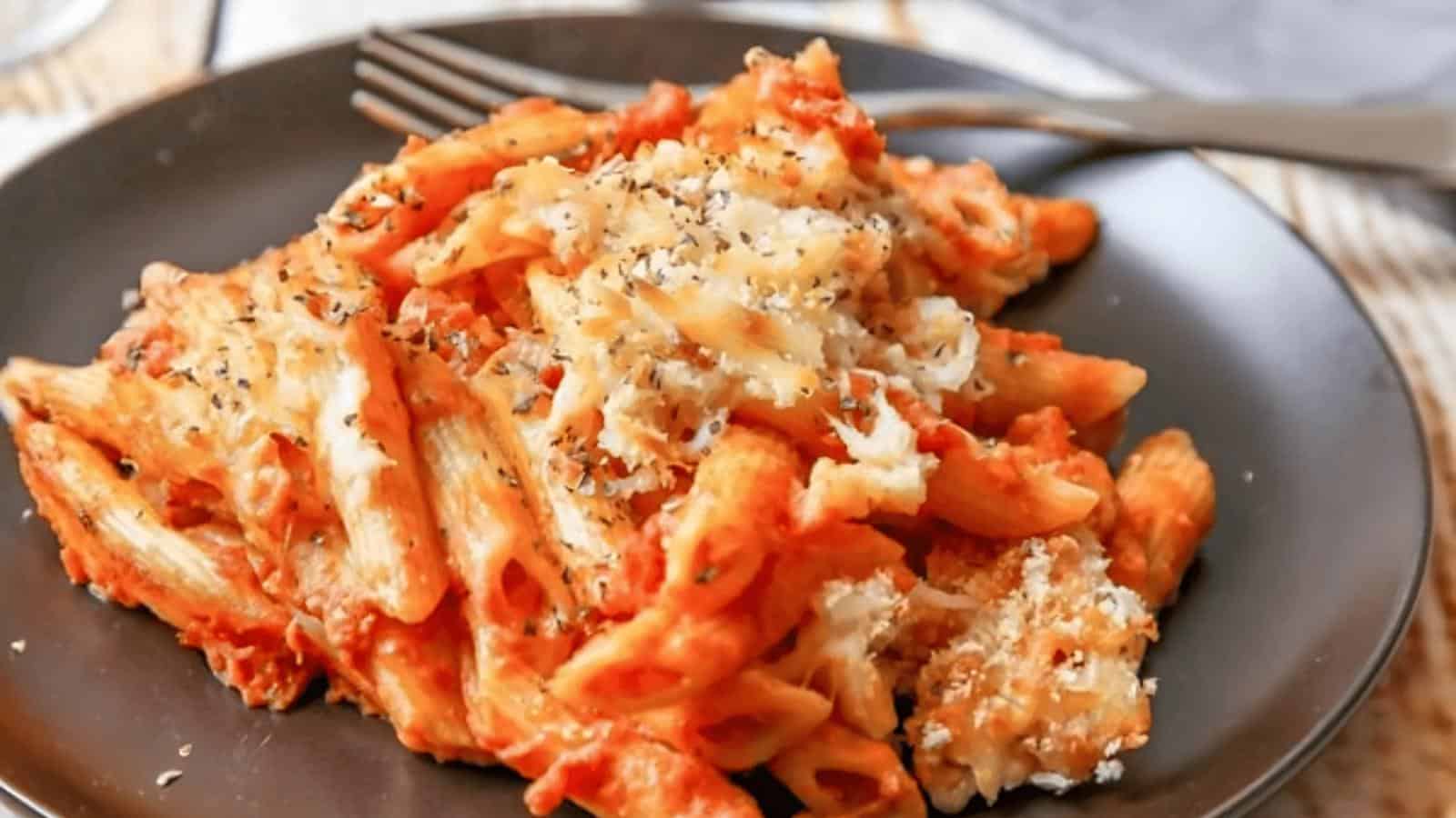A plate of baked penne pasta topped with melted cheese and herbs, with a fork placed beside it.