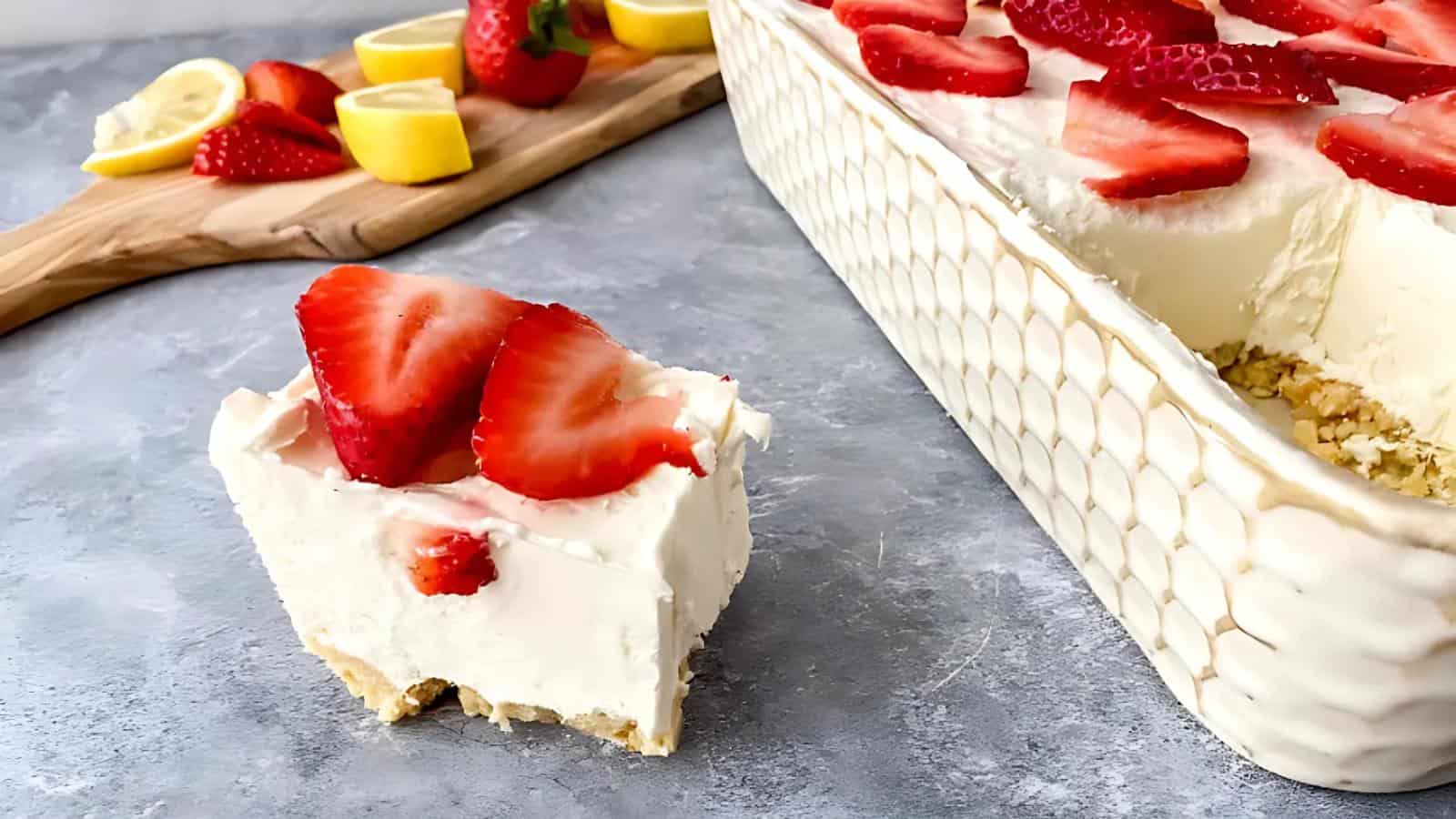 A slice of creamy cheesecake topped with sliced strawberries sits in front of a dish of the same dessert; lemons and strawberries are on a wooden board in the background.