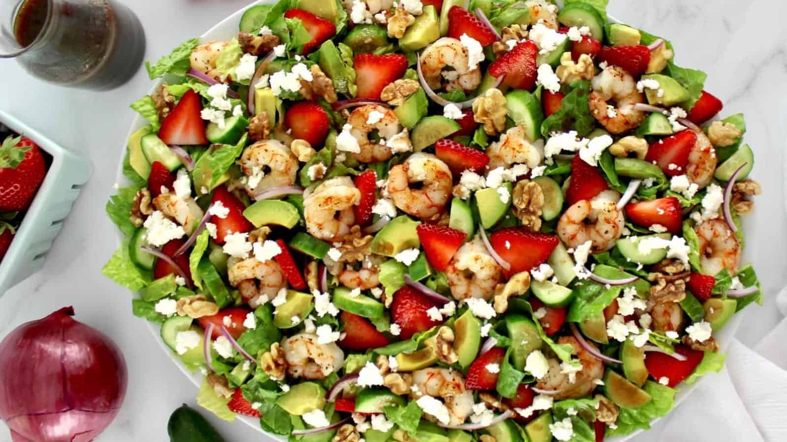 A salad with shrimp, strawberries, avocado, walnuts, red onion, spinach, and crumbled cheese arranged on a white plate. A small bottle of dressing and half a red onion are nearby.