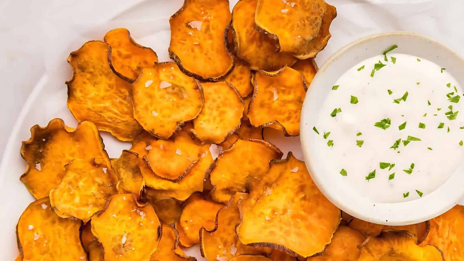 A plate of crispy sweet potato chips is served next to a bowl of creamy white dip topped with chopped herbs.