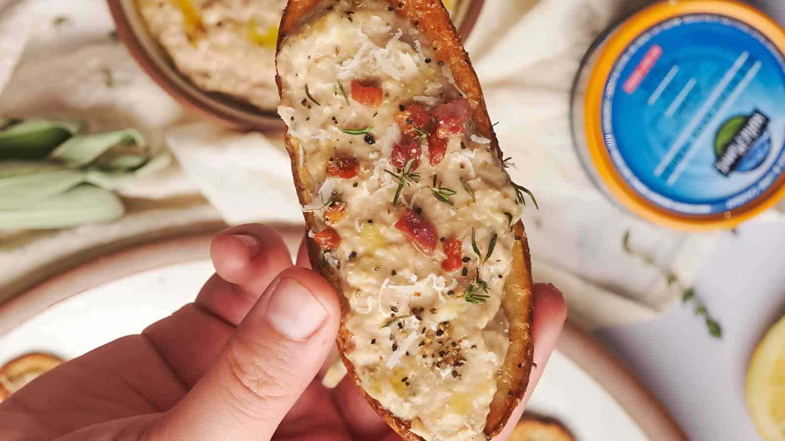 A hand holding a potato skin topped with melted cheese, herbs, and diced tomato, with a dip container and more food in the background.