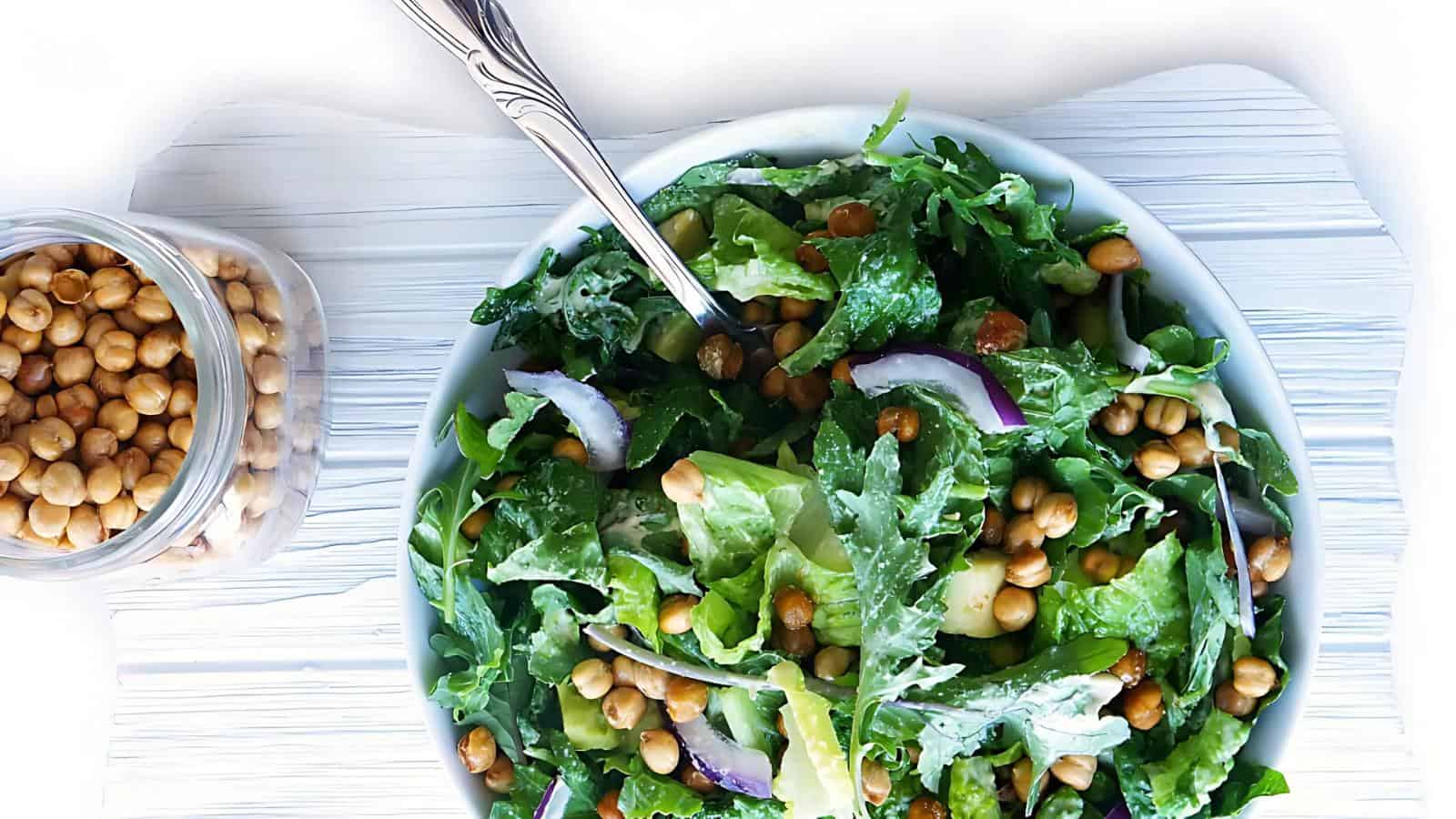 A bowl of green salad with chickpeas, red onion slices, and a creamy dressing, next to a jar of chickpeas, and a fork placed in the bowl.