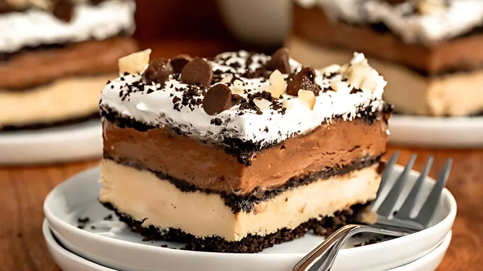 A slice of layered dessert with a chocolate topping, whipped cream, chocolate chips, and a cookie crust, served on a white plate with a fork.