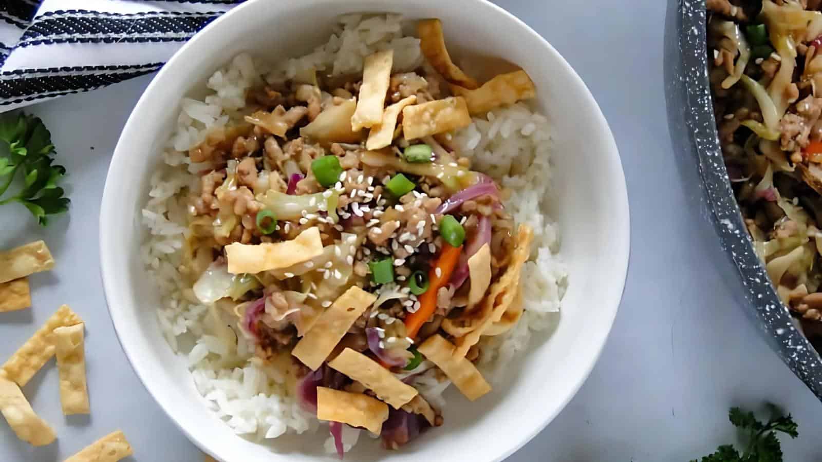 A bowl of white rice topped with stir-fried vegetables, ground meat, sesame seeds, chopped green onions, and crispy wonton strips.