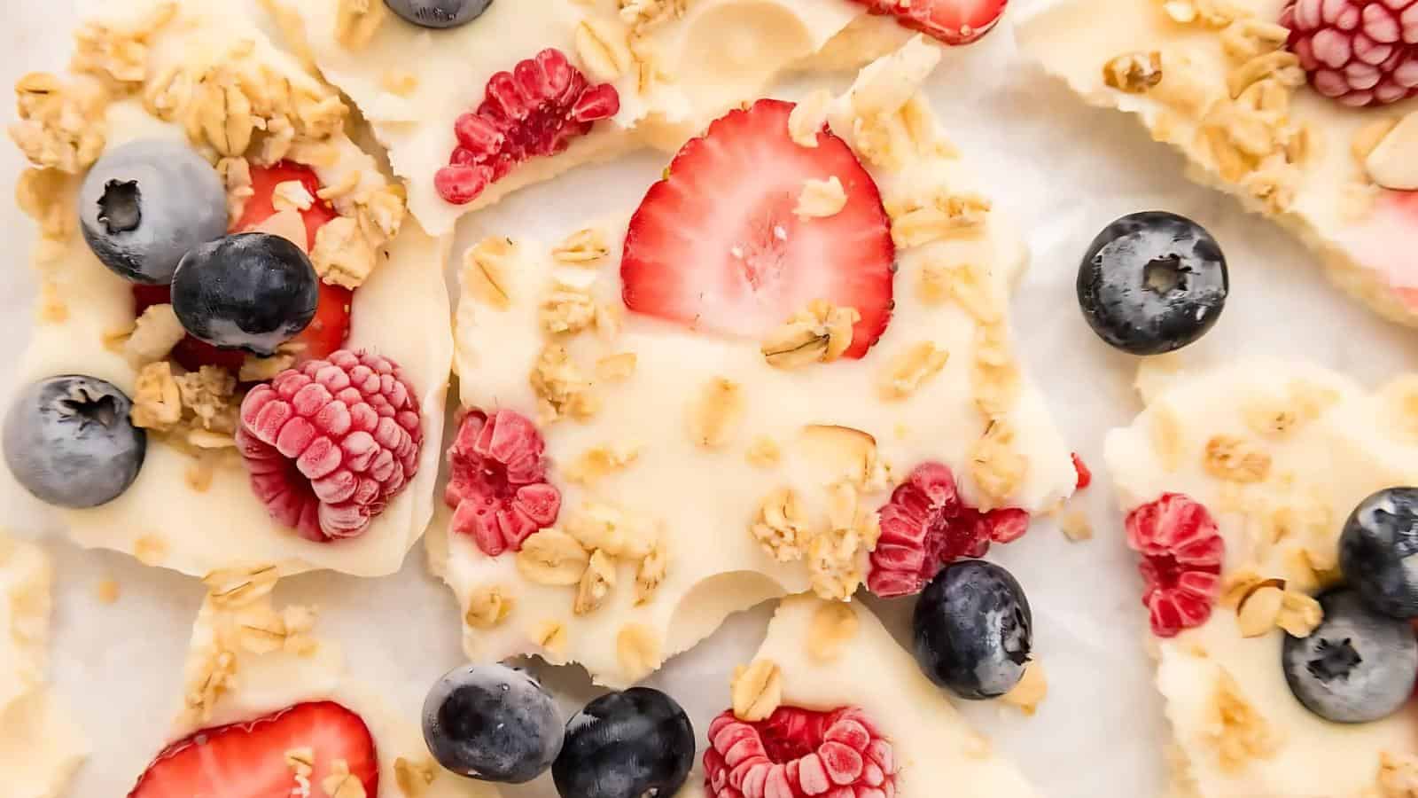 White yogurt bark topped with granola, sliced strawberries, blueberries, and raspberries, arranged on a white surface.