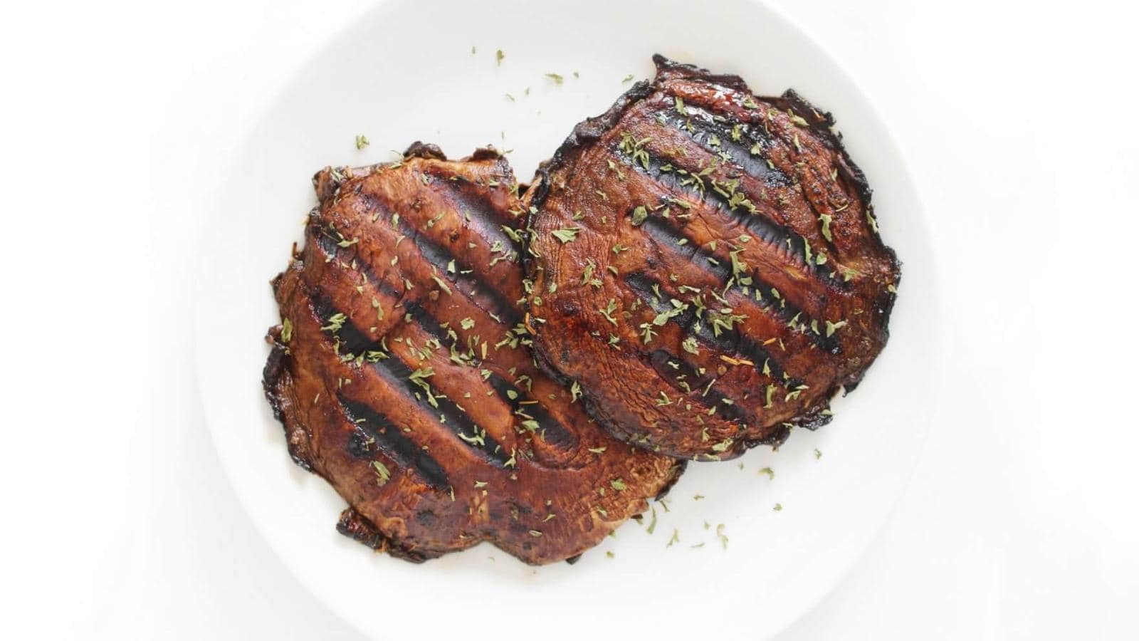 Two grilled portobello mushroom caps with visible grill marks, garnished with herbs, served on a round white plate against a white background.