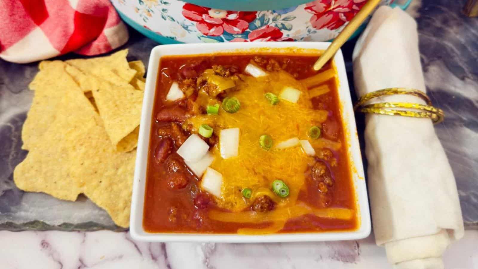 A bowl of chili topped with shredded cheese, chopped onions, and green onions, served with tortilla chips and a rolled napkin on the side.