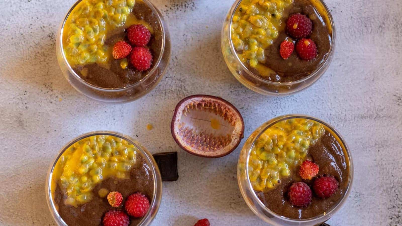 Four glasses of chocolate mousse topped with passion fruit pulp and raspberries are arranged on a light surface, with a halved passion fruit and chocolate squares in the center.