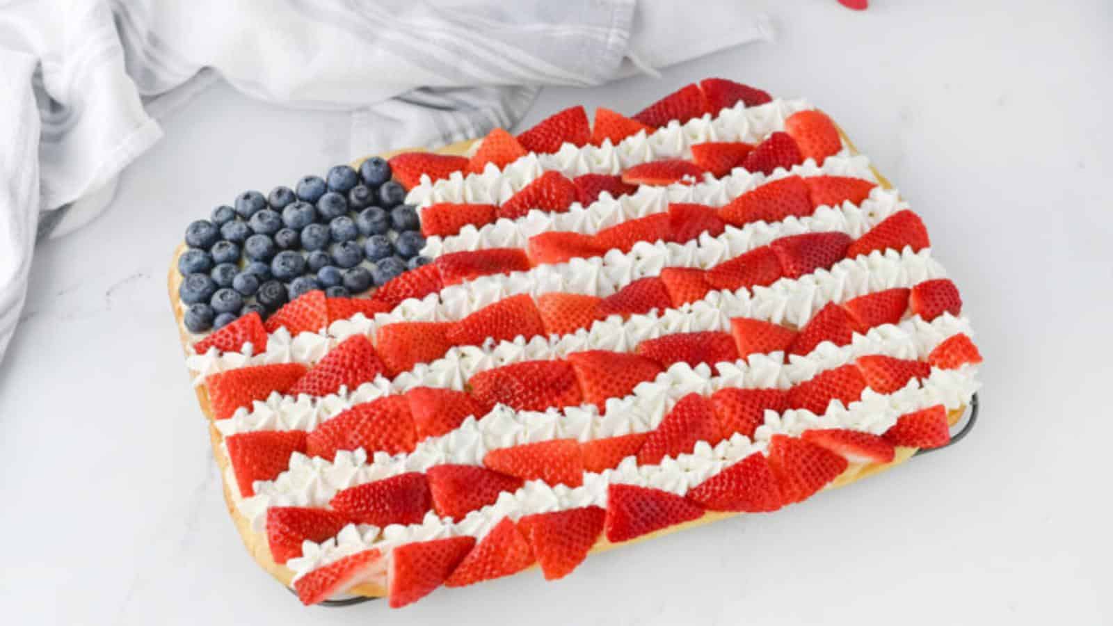 A rectangular cake decorated with blueberries, strawberries, and whipped cream to resemble the American flag, set on a white surface near a white cloth.