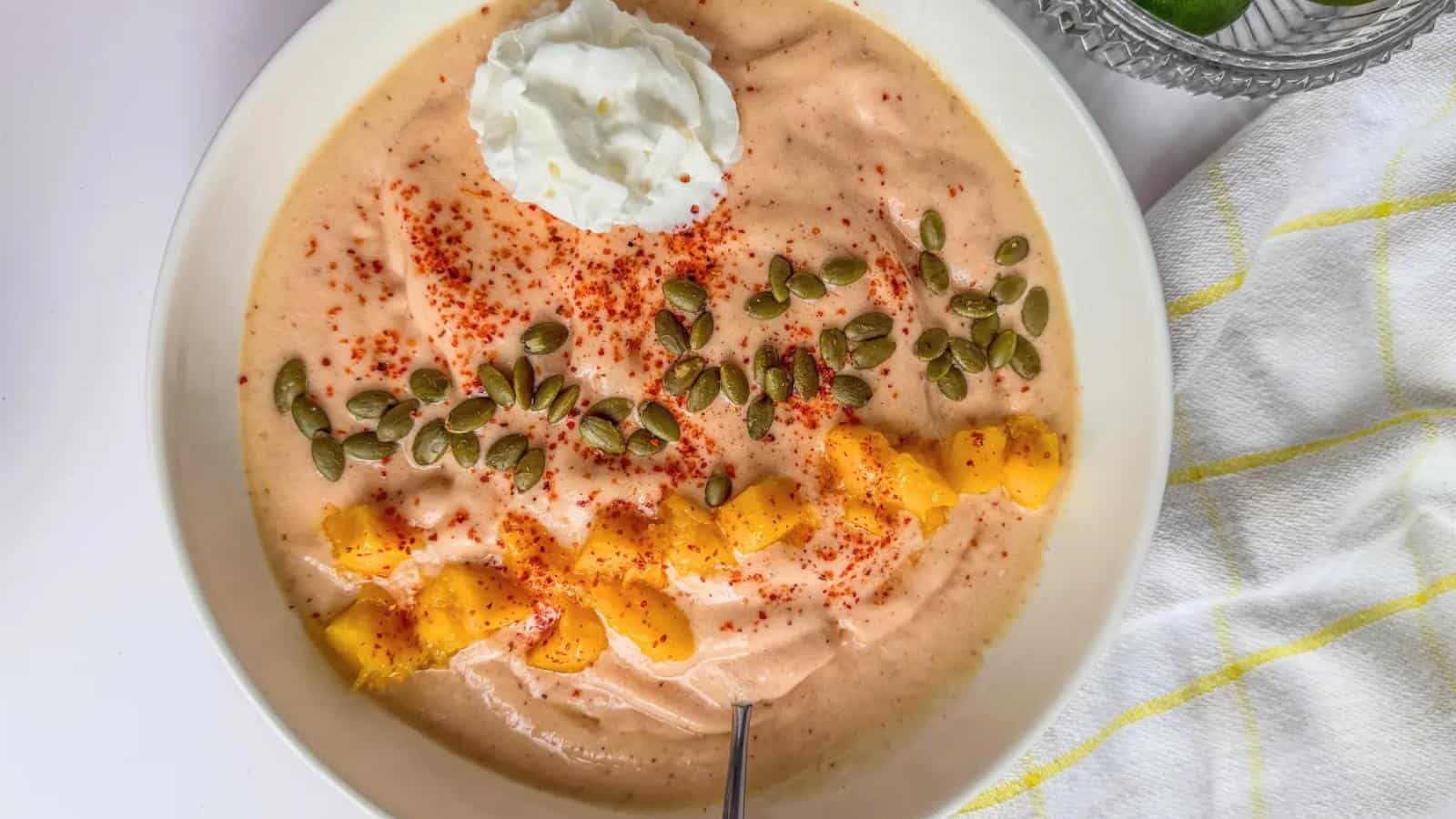 A bowl of creamy smoothie topped with pumpkin seeds, diced mango, chili powder, and a dollop of whipped cream, with a spoon resting inside.