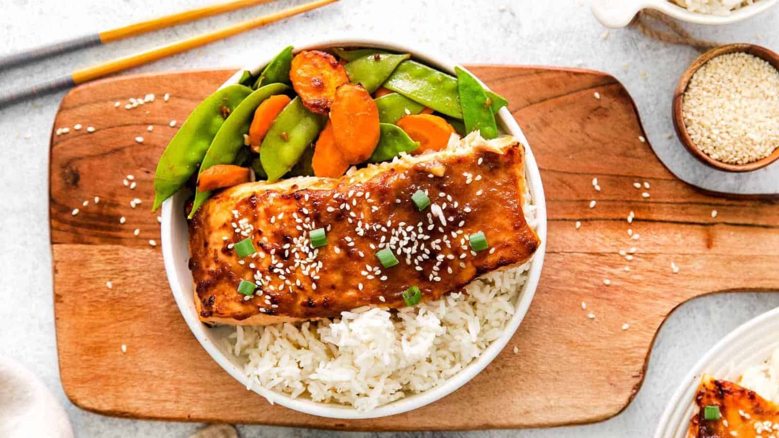 A bowl of white rice topped with glazed salmon, garnished with sesame seeds and green onions, served with sautéed snap peas and carrots.