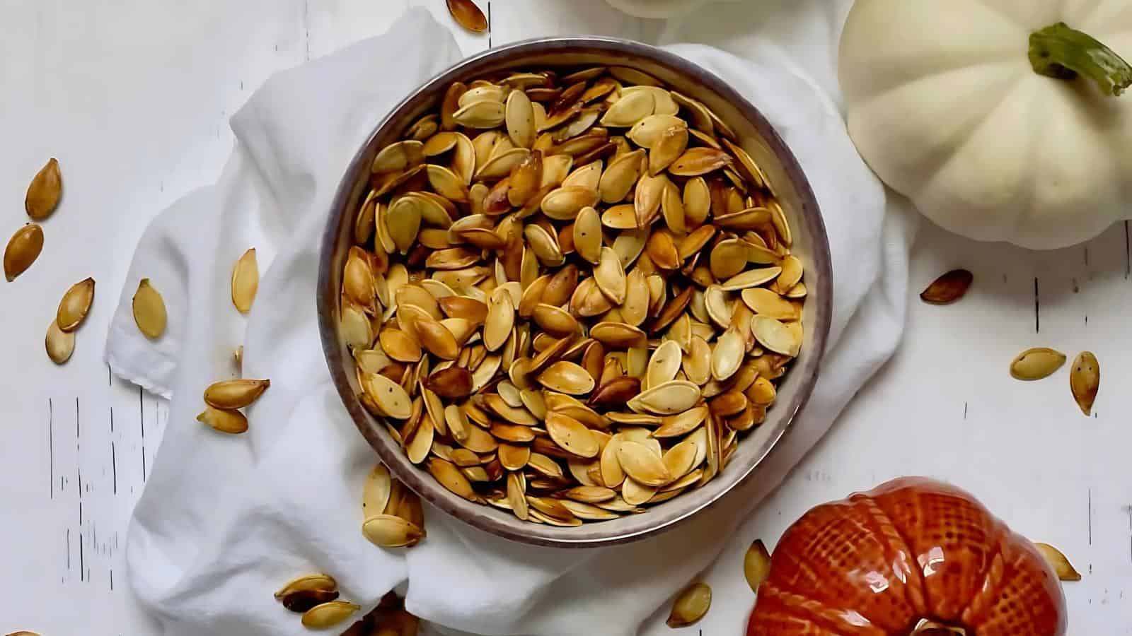 A bowl filled with roasted pumpkin seeds sits on a white cloth, with scattered seeds and two pumpkins nearby on a white surface.