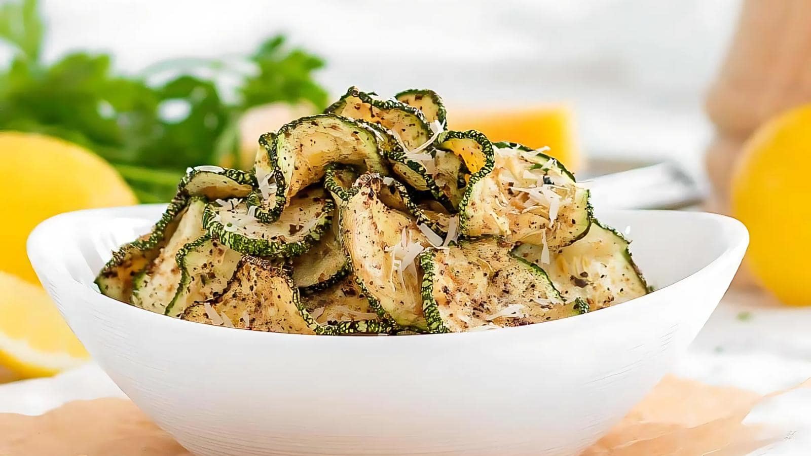 A white bowl filled with roasted zucchini slices topped with grated cheese, with lemons and herbs in the background.