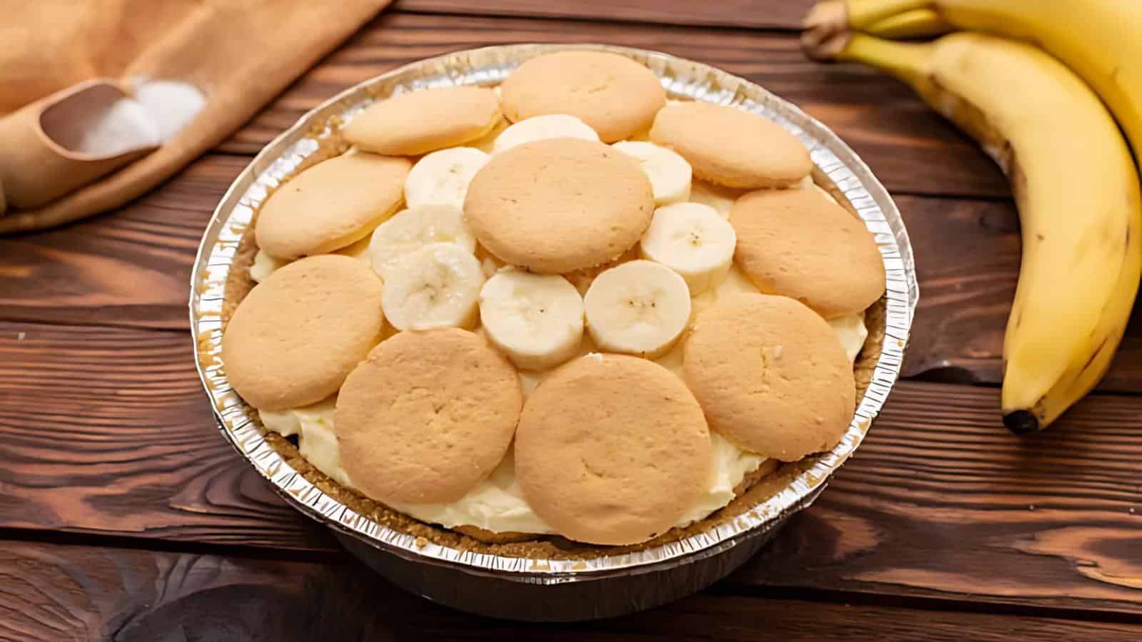 A banana pudding pie topped with round vanilla wafer cookies and banana slices, in a foil pie pan on a wooden surface next to a bunch of ripe bananas.