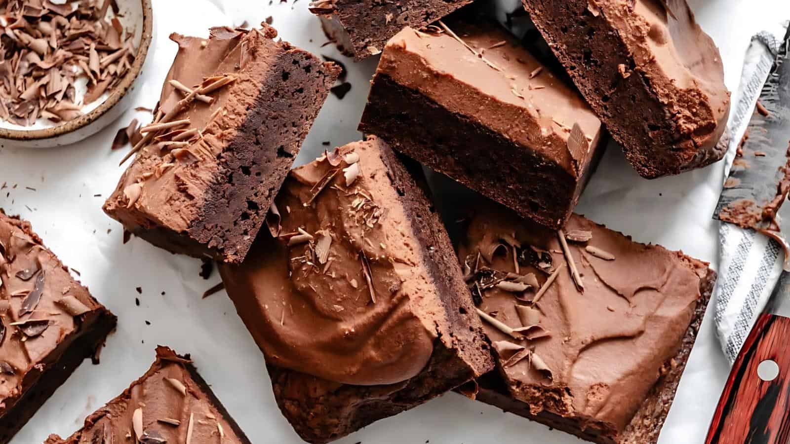 Brownies with thick chocolate frosting and chocolate shavings are arranged on a white surface next to a knife and a small bowl of chocolate.