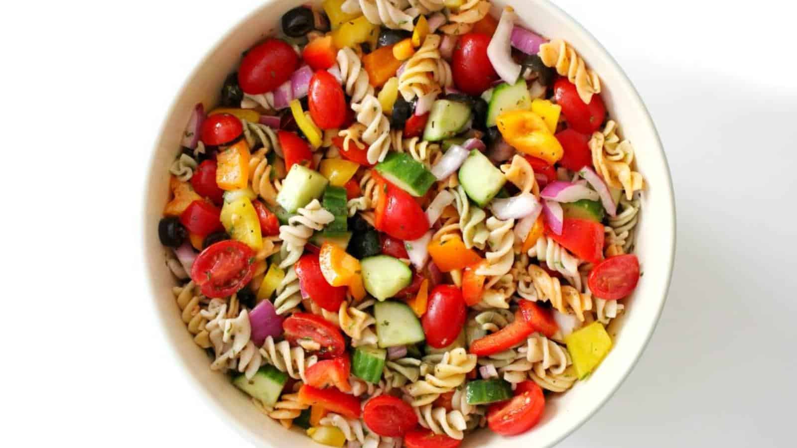 A bowl of pasta salad with rotini, cherry tomatoes, cucumbers, bell peppers, red onions, and black olives, mixed together on a white background.