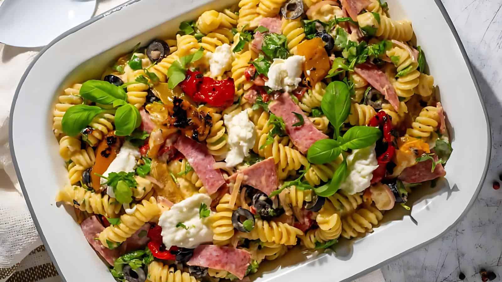 A white dish filled with rotini pasta, sliced ham, black olives, roasted peppers, fresh basil leaves, and dollops of cheese.