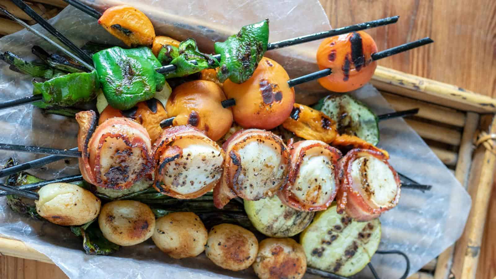 Skewers with grilled vegetables, tomatoes, peppers, and scallops wrapped in bacon, served on parchment paper over a wooden tray.