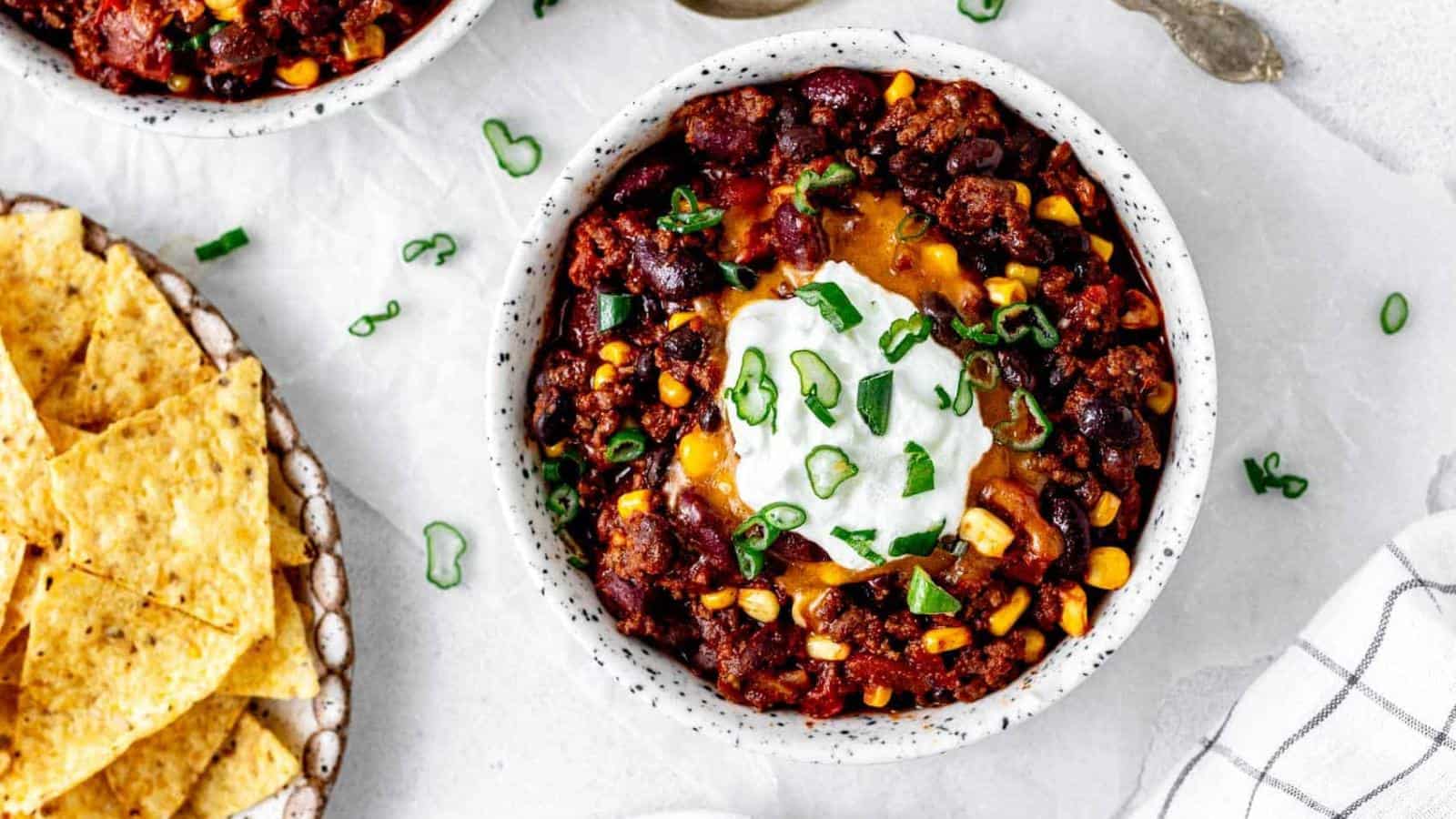 A bowl of chili topped with sour cream, shredded cheese, and chopped green onions, served next to a plate of tortilla chips.