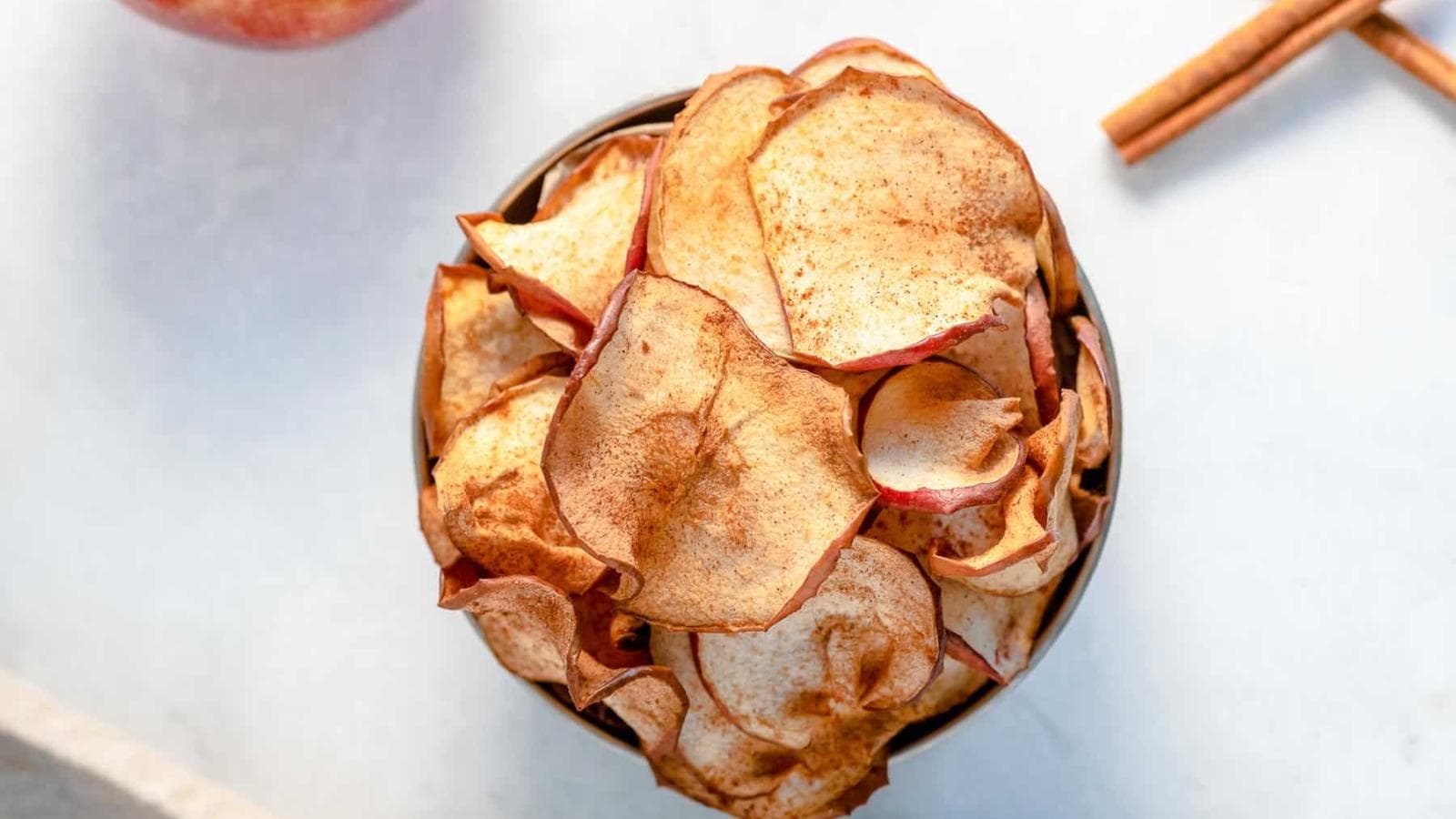 A bowl filled with thinly sliced, baked apple chips, lightly dusted with cinnamon, is shown on a light surface with a cinnamon stick and part of an apple nearby.