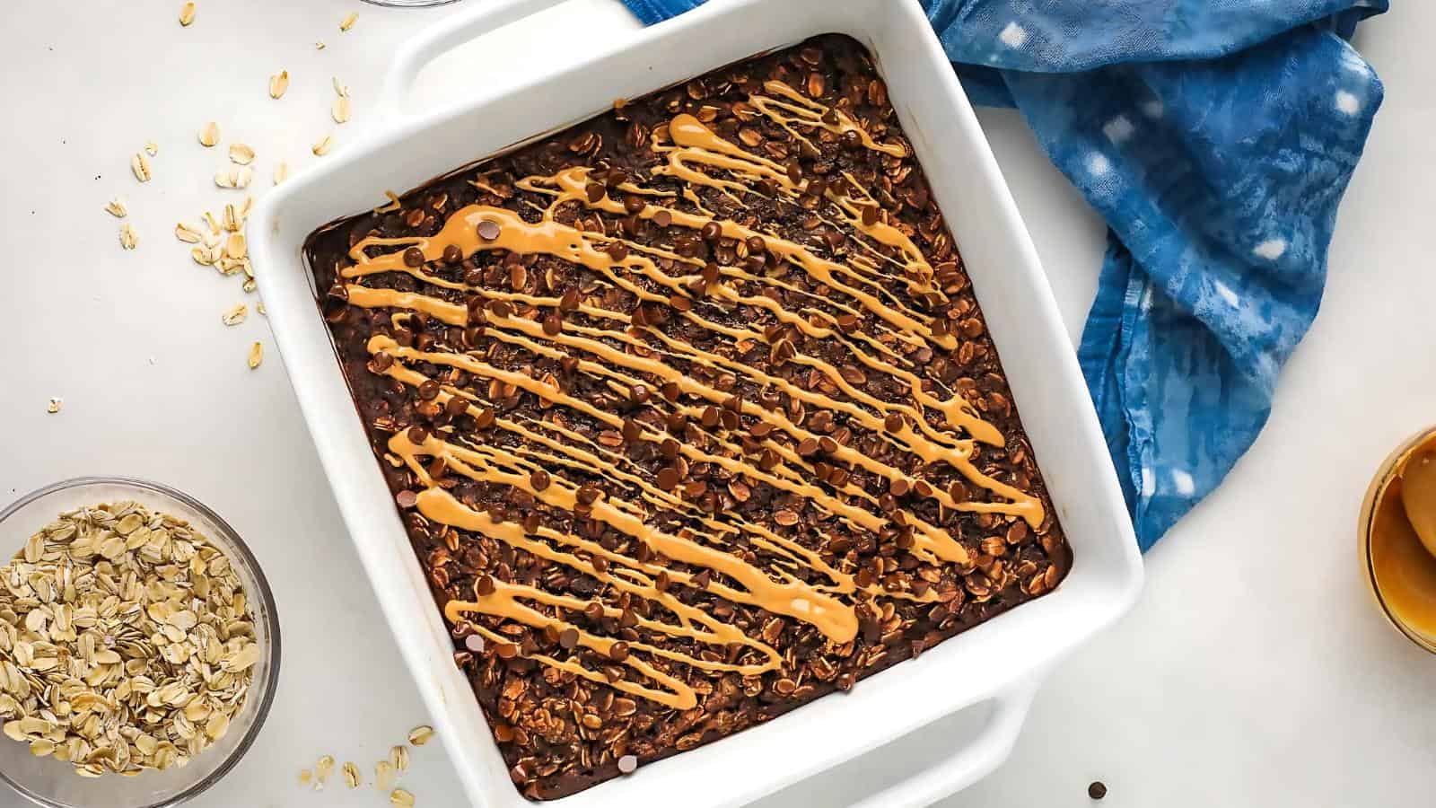 A baked oatmeal dish topped with a drizzle of peanut butter sits in a white square pan, with oats, a blue cloth, and a bowl of honey nearby.