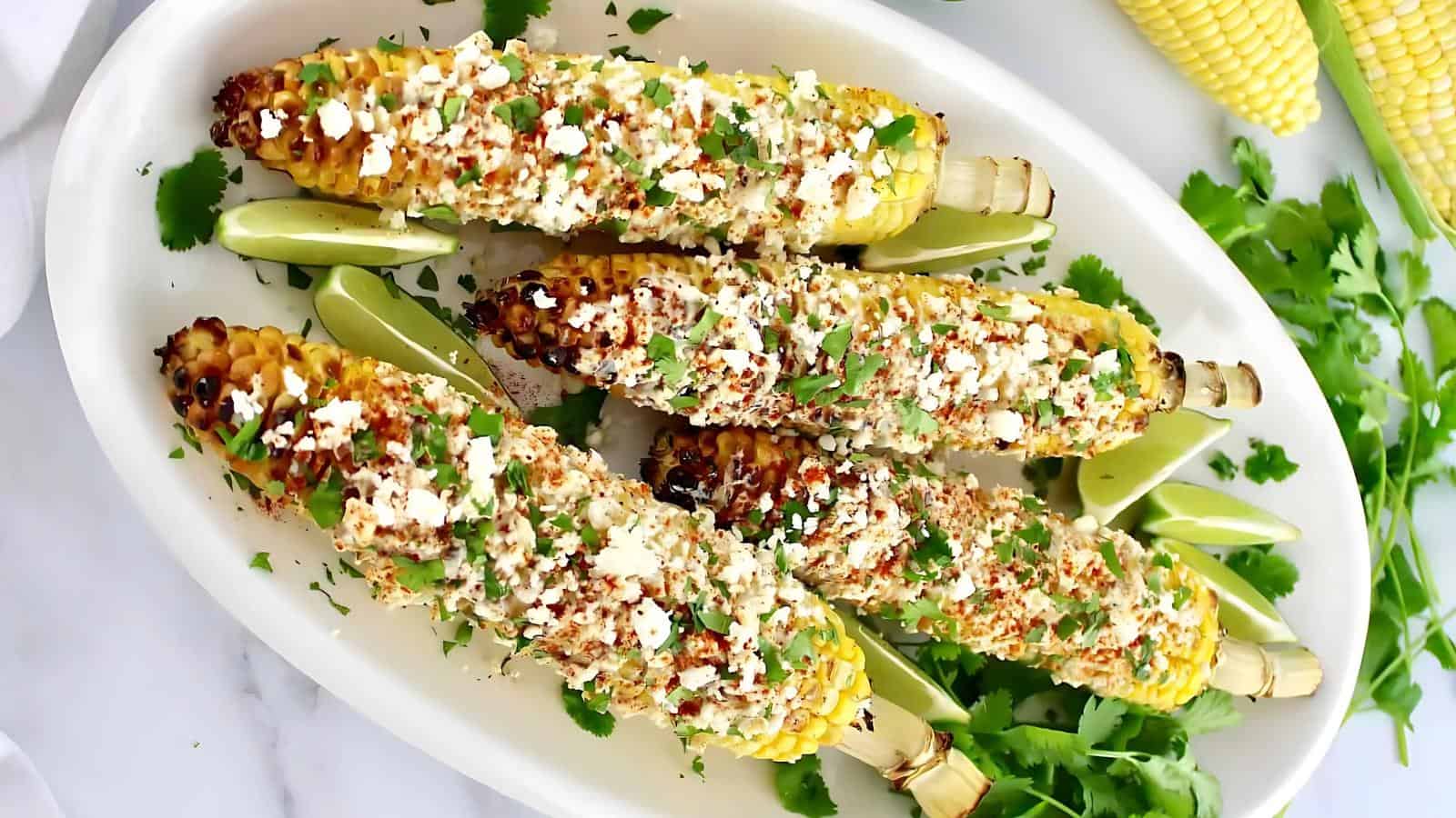 Grilled corn on the cob topped with cheese, spices, and chopped cilantro, served on a white platter with lime wedges and fresh cilantro.