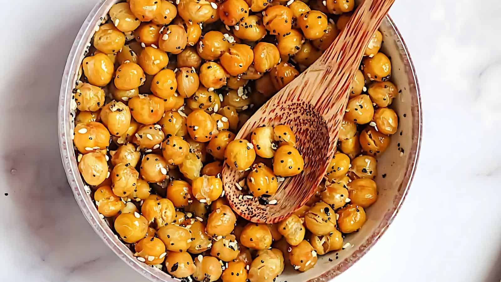 A bowl of roasted chickpeas topped with sesame seeds and black pepper, with a wooden spoon resting inside the bowl on a white surface.