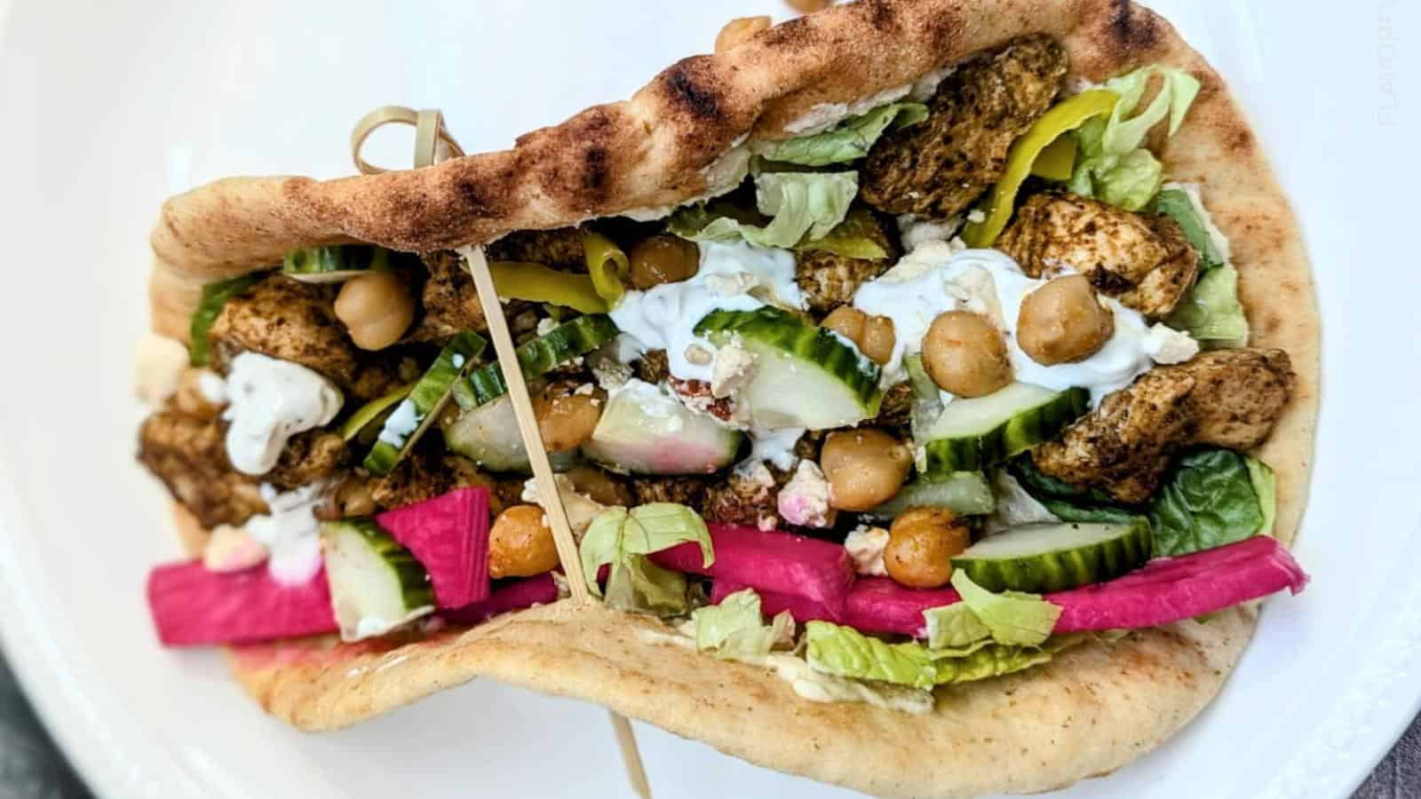 A pita sandwich filled with grilled chicken, chickpeas, lettuce, cucumber, pickled vegetables, tzatziki sauce, and feta cheese on a white plate.