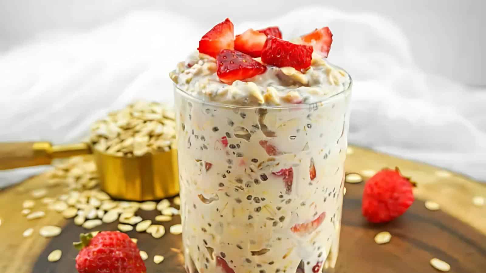 A glass filled with overnight oats mixed with chia seeds and strawberries, topped with strawberry pieces, sits on a wooden surface with loose oats and whole strawberries nearby.