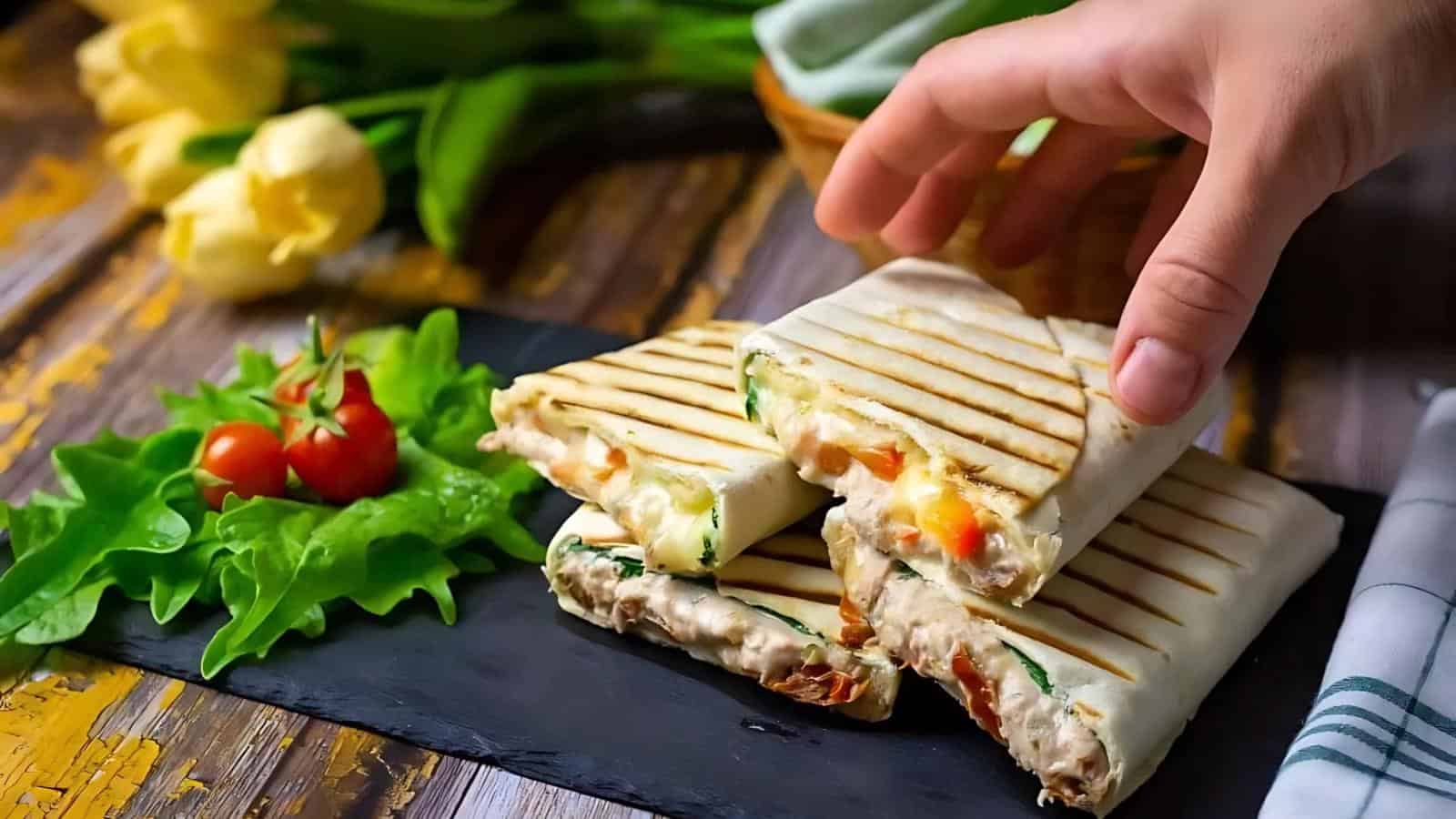 A hand reaches for a grilled wrap filled with vegetables and cheese, placed on a slate board with lettuce and cherry tomatoes, with yellow flowers in the background.