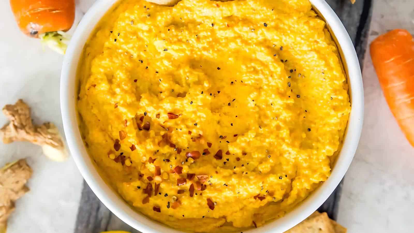 A bowl of creamy orange hummus, garnished with red pepper flakes and black pepper, surrounded by carrot slices and crackers.