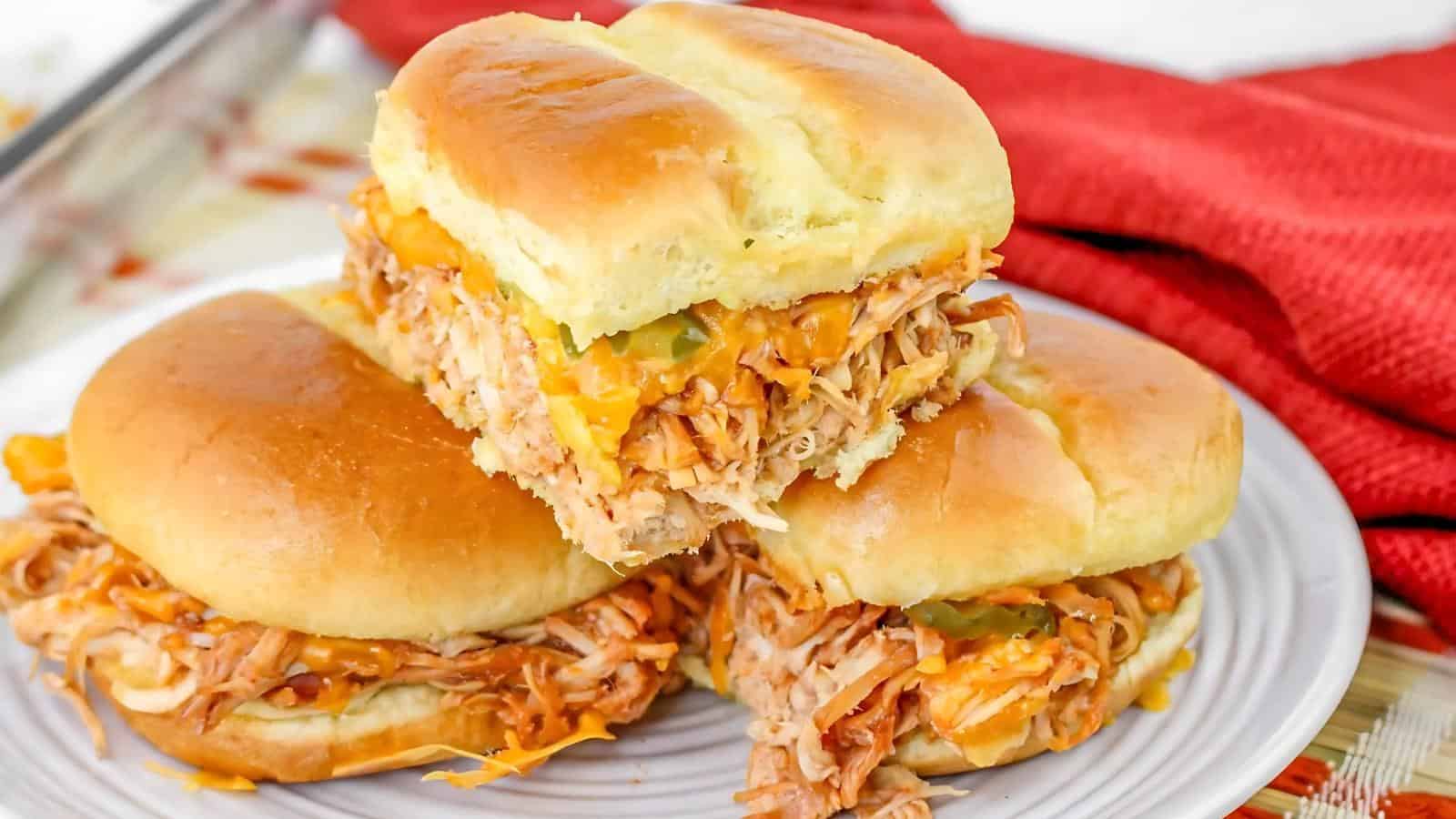 Three pulled chicken sandwiches with melted cheese and pickles on soft buns are stacked on a white plate, with a red cloth in the background.
