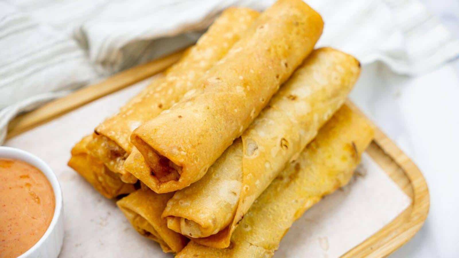 A stack of crispy, golden-brown rolled taquitos on a tray with a small bowl of orange dipping sauce on the side.