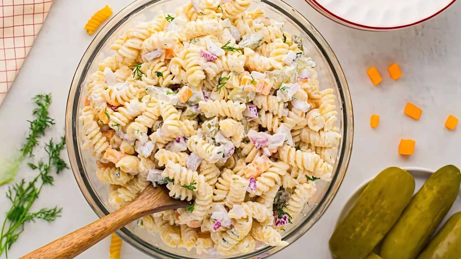 A bowl of creamy pasta salad with rotini, diced red onions, pickles, and herbs, with a wooden spoon, next to sliced pickles and small cheese cubes on a white surface.