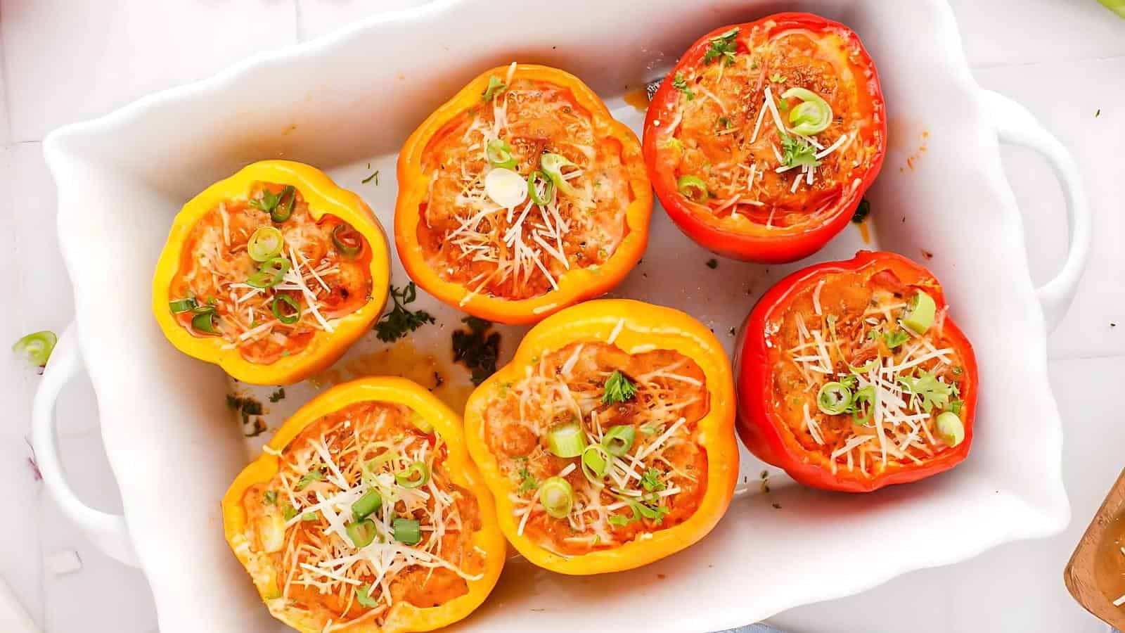 Six stuffed bell peppers, red and yellow, filled with a cheesy tomato mixture and topped with sliced green onions, are arranged in a white baking dish.