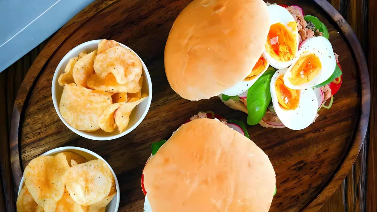 Two sandwiches with sliced boiled eggs and vegetables on a wooden tray, served with two small bowls of potato chips.