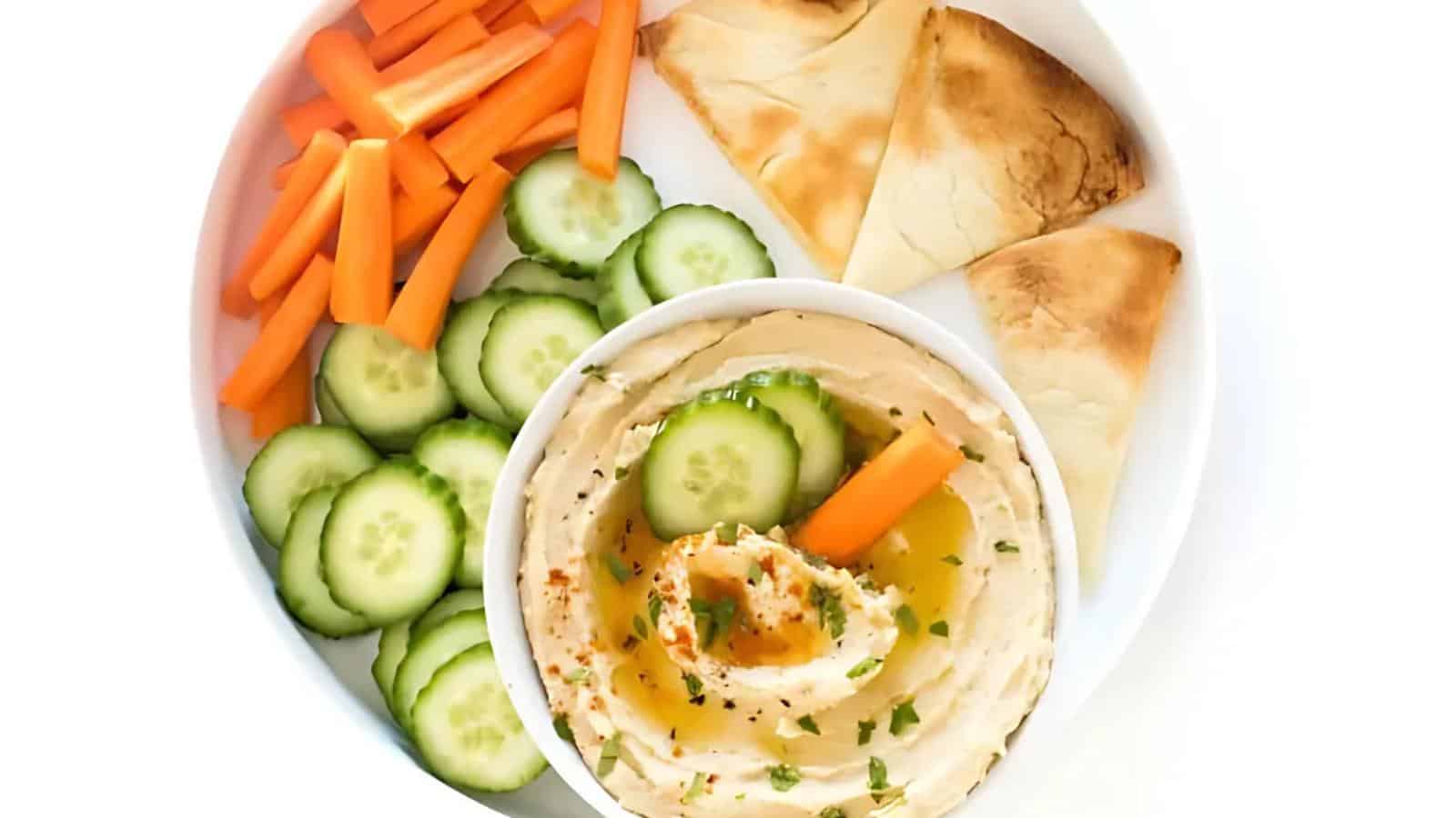 A plate with carrot sticks, cucumber slices, pita bread pieces, and a bowl of hummus topped with olive oil and herbs.
