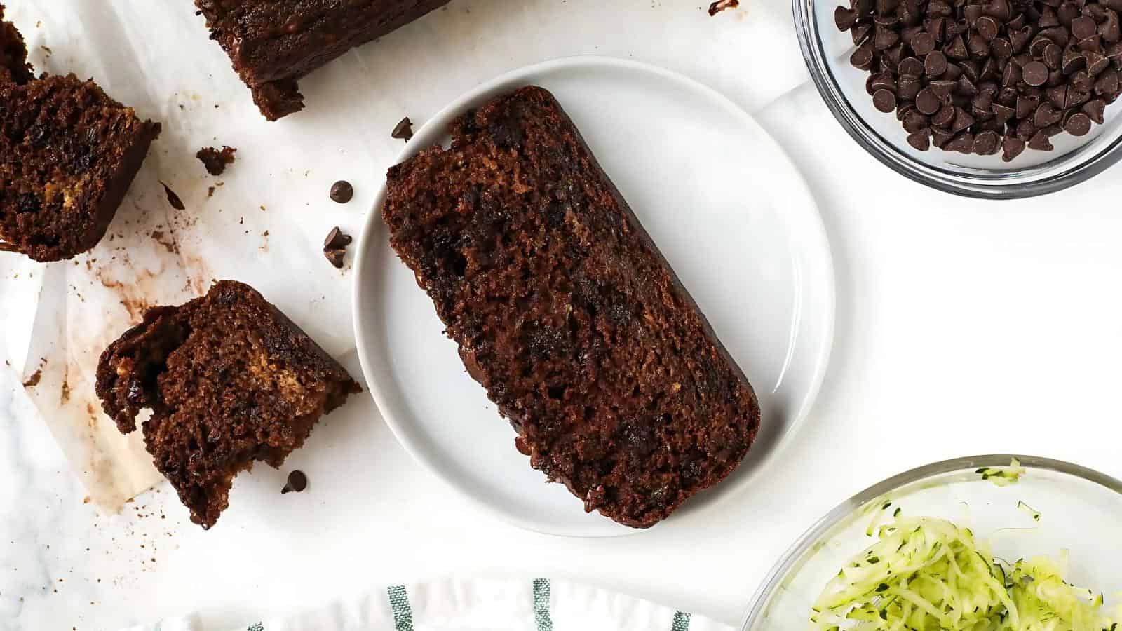 A slice of chocolate zucchini bread on a white plate, surrounded by chocolate chips, shredded zucchini, and additional slices of bread.
