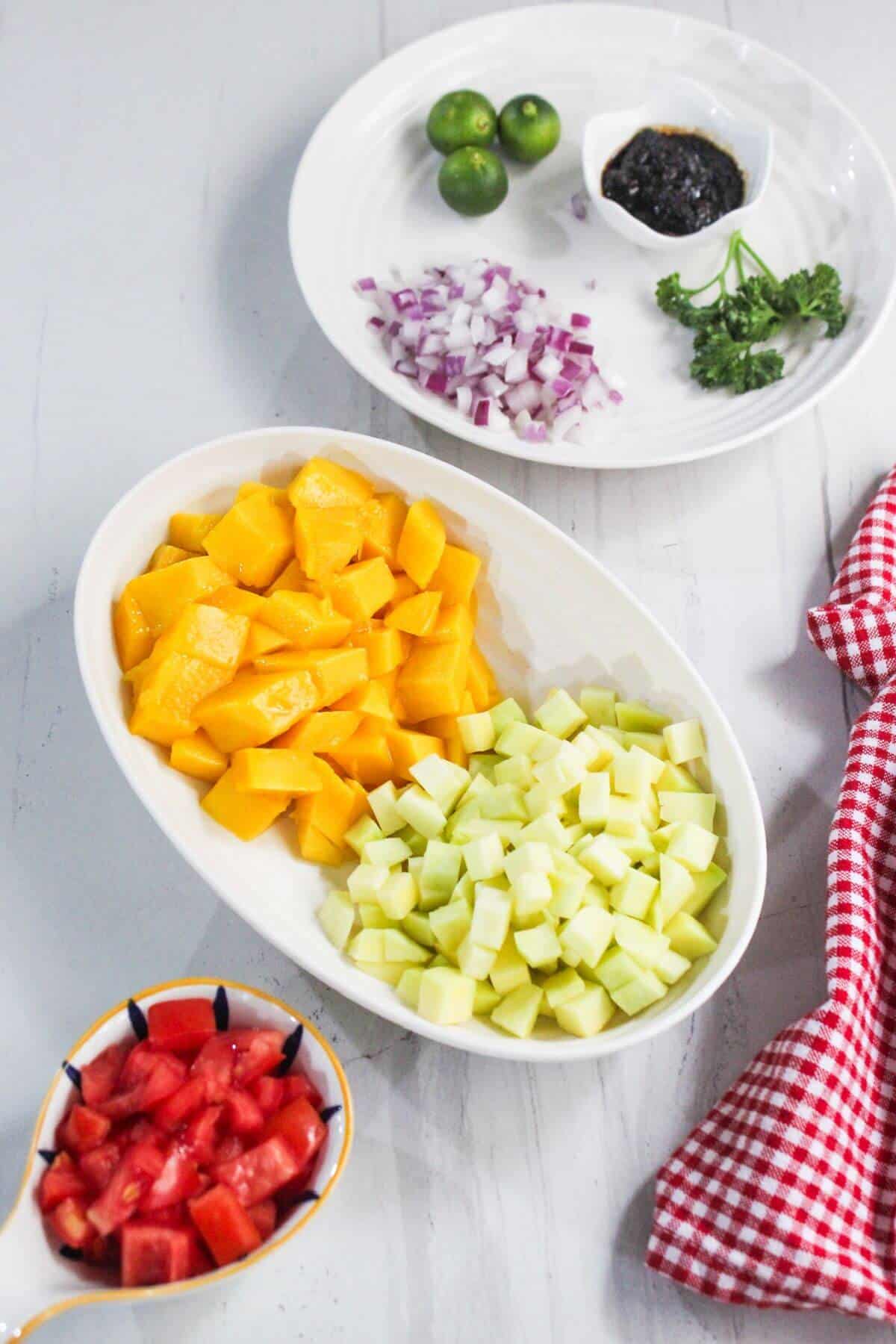 Chopped mango, cucumber, and tomato in bowls with diced red onion, calamansi, parsley, and a dark sauce on a white plate; a red checked cloth on the side.